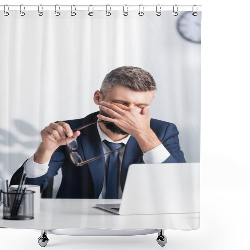 Personality  Tired businessman holding eyeglasses and touching eyes near laptop on blurred foreground  shower curtains